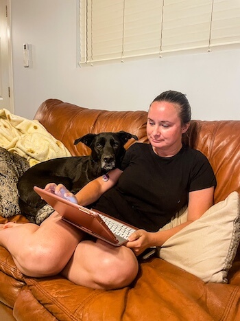 A woman reads on her laptop with her dog looking at the camera.