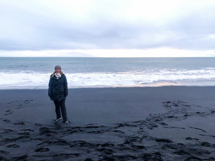 A woman stands on a black sand beach in Iceland.
